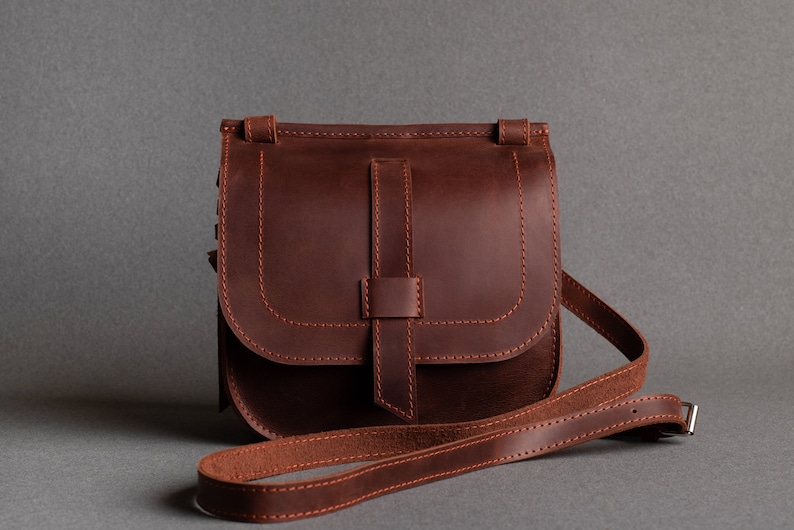 May include: A brown leather crossbody bag with a flap closure and an adjustable strap. The bag has a stitched design and a simple, classic style.