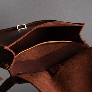 May include: A brown leather bag with a flap top and a buckle closure. The bag is open and the inside is lined with a brown fabric.