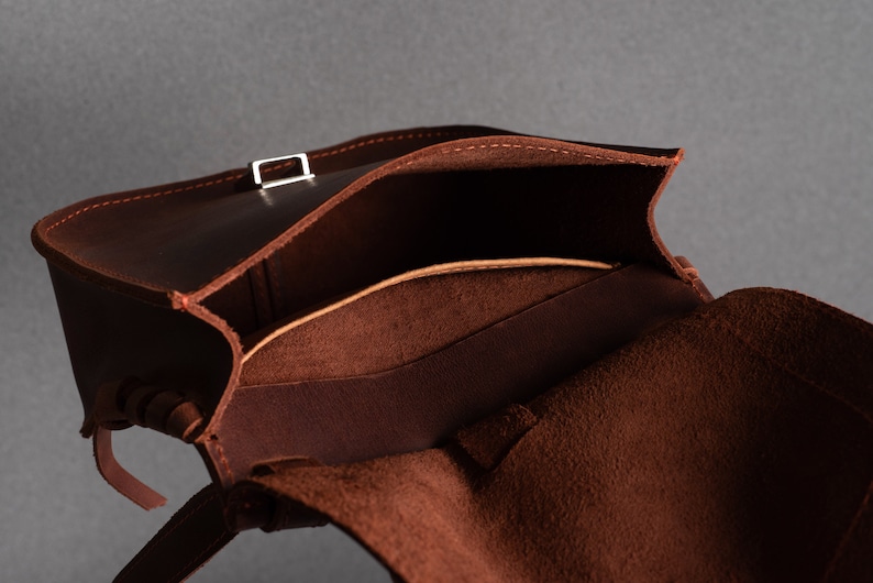 May include: A brown leather bag with a flap top and a buckle closure. The bag is open and the inside is lined with a brown fabric.