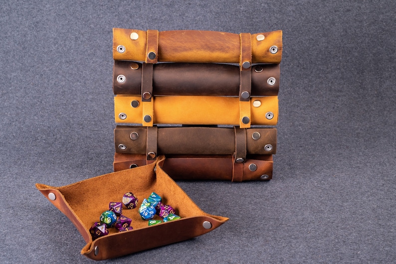 May include: A brown leather dice tray with a set of colorful dice. The tray is square and has a rolled edge. The dice are scattered inside the tray. There is a stack of five brown leather rolls with metal snaps on the side.