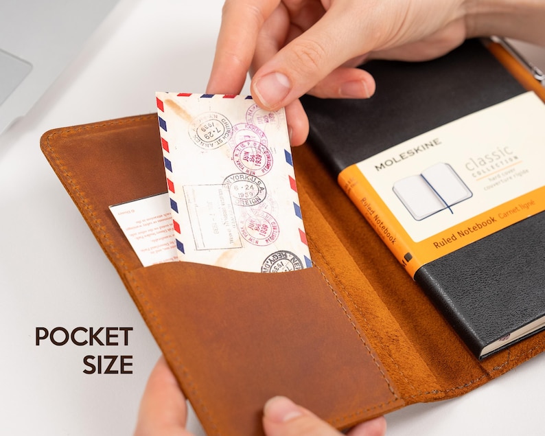 Pode incluir: Uma capa de caderno de bolso em couro castanho com um caderno Moleskine preto e um envelope de correio a&eacute;reo vintage. A capa tem um bolso que cont&eacute;m o envelope e um pequeno cart&atilde;o. O texto "POCKET SIZE" &eacute; vis&iacute;vel.