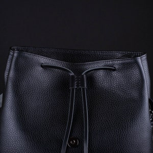 May include: A black leather bucket bag with a drawstring closure and a small metal button. The bag has a textured surface and a strap.