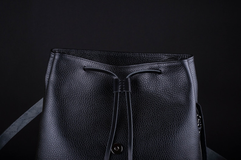 May include: A black leather bucket bag with a drawstring closure and a small metal button. The bag has a textured surface and a strap.