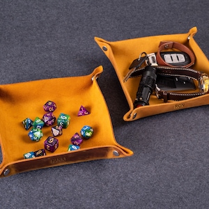 May include: Two brown leather catchall trays with personalized names. One tray has a collection of colorful dice, the other has a watch, keys, and a flashlight.