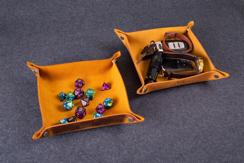 May include: Two brown leather catchall trays with personalized names. One tray has a collection of colorful dice, the other has a watch, keys, and a flashlight.