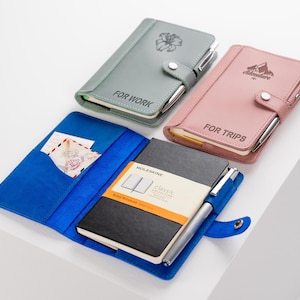 Pode incluir: Uma cole&ccedil;&atilde;o de cadernos em v&aacute;rias cores, incluindo cinza, rosa e azul. Os cadernos t&ecirc;m porta-canetas e s&atilde;o rotulados como "FOR WORK" e "FOR TRIPS". Um caderno est&aacute; aberto, revelando um caderno Moleskine preto e um bolso com cart&otilde;es.
