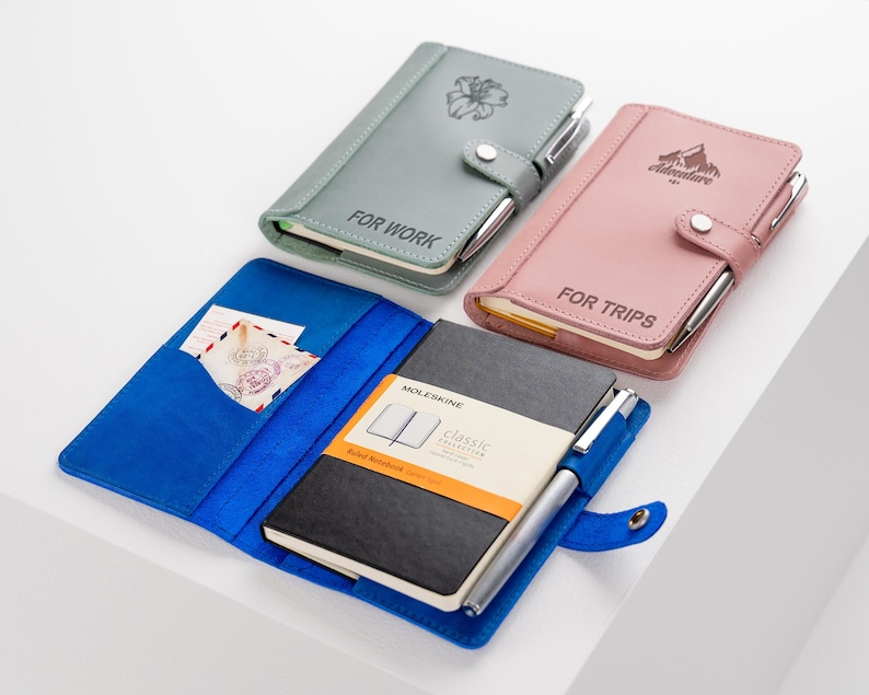 Pode incluir: Uma cole&ccedil;&atilde;o de cadernos em v&aacute;rias cores, incluindo cinza, rosa e azul. Os cadernos t&ecirc;m porta-canetas e s&atilde;o rotulados como "FOR WORK" e "FOR TRIPS". Um caderno est&aacute; aberto, revelando um caderno Moleskine preto e um bolso com cart&otilde;es.