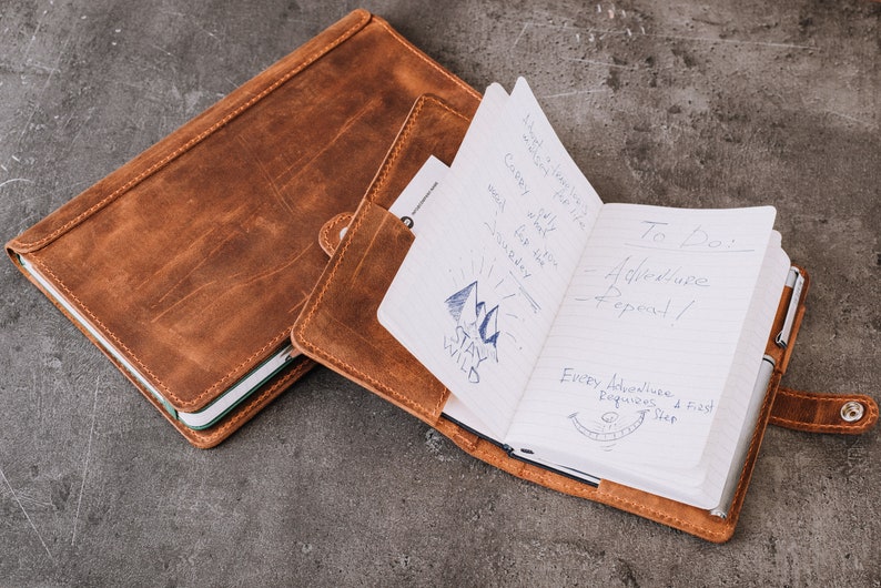 Pode incluir: Um di&aacute;rio de couro castanho com fecho de press&atilde;o, aberto para revelar notas manuscritas e uma caneta. As p&aacute;ginas do di&aacute;rio exibem o texto "To Do: Adventure - Repeat!" e um gr&aacute;fico de montanha com as palavras "Stay Wild".
