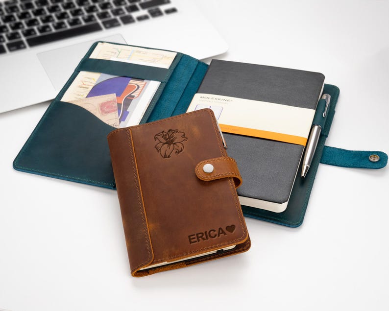 Pode incluir: Uma cole&ccedil;&atilde;o de cadernos de couro e uma caneta sobre uma superf&iacute;cie branca. Um caderno de couro castanho com um desenho floral e o nome "ERICA" em primeiro plano. Um caderno cinzento escuro com uma faixa branca e laranja atr&aacute;s. Um caderno de couro verde-azulado est&aacute; aberto.