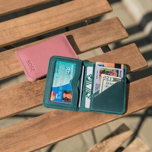 May include: Two leather wallets, one pink and one teal, with the word "ROSE" embossed on the pink wallet. The teal wallet is open and has several cards and cash inside.
