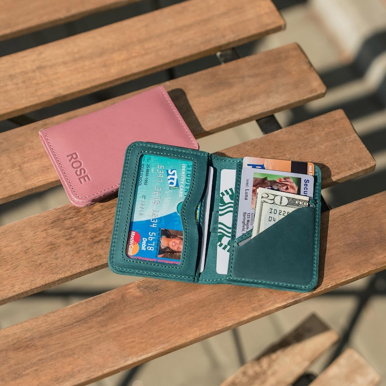 May include: Two leather wallets, one pink and one teal, with the word "ROSE" embossed on the pink wallet. The teal wallet is open and has several cards and cash inside.