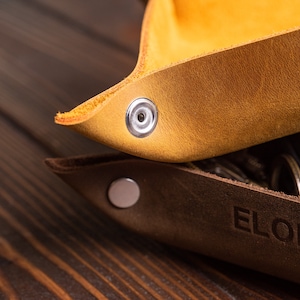 May include: Two leather trays, one brown and one yellow, with metal snaps. The brown tray has the text "ELON MU" embossed on it.