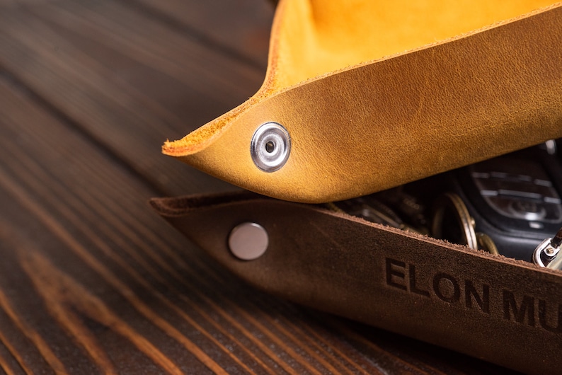May include: Two leather trays, one brown and one yellow, with metal snaps. The brown tray has the text "ELON MU" embossed on it.
