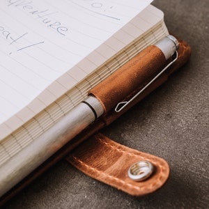 Pode incluir: Close-up de um caderno com capa de couro castanho e uma caneta prateada num porta-canetas de couro a condizer. O caderno est&aacute; aberto, revelando papel pautado com texto manuscrito. O porta-canetas est&aacute; preso &agrave; capa.