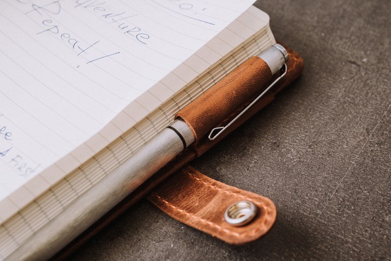 Pode incluir: Close-up de um caderno com capa de couro castanho e uma caneta prateada num porta-canetas de couro a condizer. O caderno est&aacute; aberto, revelando papel pautado com texto manuscrito. O porta-canetas est&aacute; preso &agrave; capa.