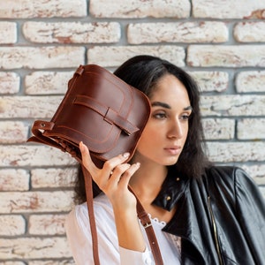 May include: A brown leather crossbody bag with a flap closure and a strap. The bag is held by a person wearing a white shirt and a black leather jacket.