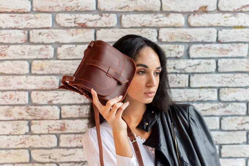 May include: A brown leather crossbody bag with a flap closure and a strap. The bag is held by a person wearing a white shirt and a black leather jacket.