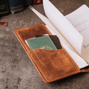 Pode incluir: Um di&aacute;rio de couro castanho com um bolso contendo pequenos cart&otilde;es e um caderno com anota&ccedil;&otilde;es manuscritas. O di&aacute;rio est&aacute; aberto, revelando p&aacute;ginas pautadas. O texto no caderno diz "To Do: Adventure repeat!" e "Every Adventure Requires Sleep".