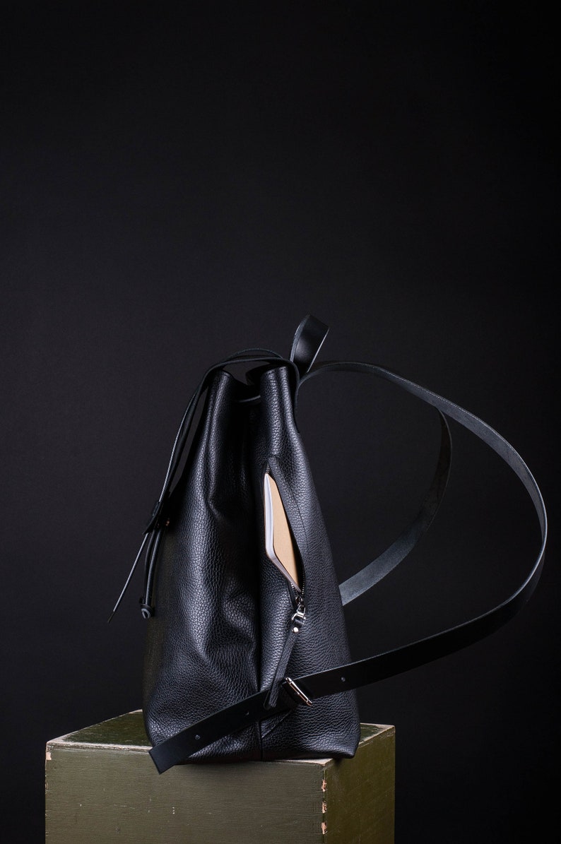 May include: A black leather backpack with a zippered pocket on the side. The backpack is sitting on a green surface.