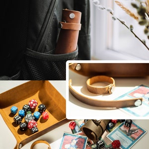 May include: A brown leather dice tray with a rolled up leather dice bag and a set of dice. The tray has a snap closure and a leather strap. The dice bag has a snap closure and a leather strap. The dice are a variety of colors and sizes. The dice tray and bag are on a white surface with a few playing cards.