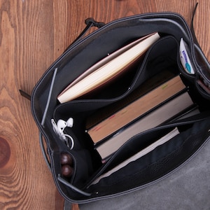 May include: A black leather backpack with multiple compartments. The backpack is open and the interior is lined with black fabric. The backpack contains a book, a notebook, a phone, and a pair of headphones.