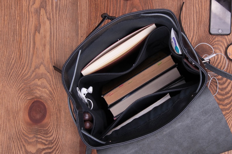 May include: A black leather backpack with multiple compartments. The backpack is open and the interior is lined with black fabric. The backpack contains a book, a notebook, a phone, and a pair of headphones.