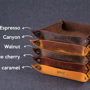 May include: A stack of six leather trays in various shades of brown, including espresso, canyon, walnut, antique cherry, and brown caramel. Each tray has a name embossed on it: "Marry", "Chris", "Elizabeth", "Katherine", and "Mike".