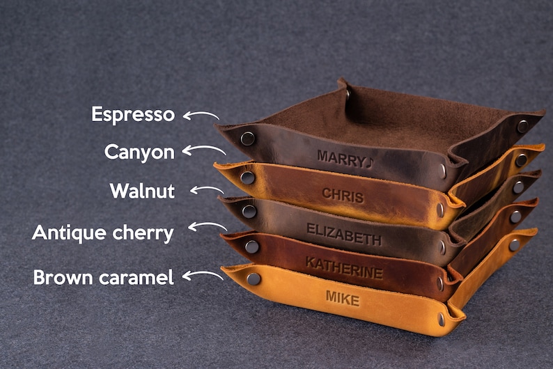 May include: A stack of six leather trays in various shades of brown, including espresso, canyon, walnut, antique cherry, and brown caramel. Each tray has a name embossed on it: "Marry", "Chris", "Elizabeth", "Katherine", and "Mike".
