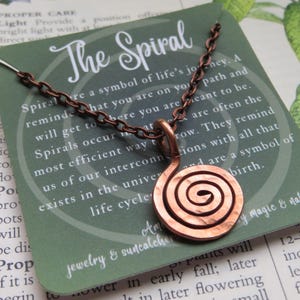 May include: A copper-colored spiral pendant necklace. The pendant is a hammered spiral design, suspended from a matching chain. The necklace is displayed on a green card with the text "The Spiral".