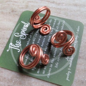 May include: Three copper wire rings, each featuring a double spiral design. The rings are displayed on a square green card with white text that reads The Spiral and includes descriptive text about the symbol of life and nature.
