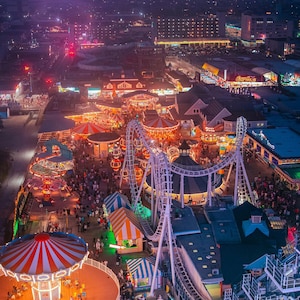 An aerial view of an amusement park at night. The park is lit up with colorful lights, and there are many rides and attractions visible, including a carousel, a roller coaster, and a Ferris wheel. The park is located on a boardwalk, and there are many people walking around and enjoying the rides. The sky is a dark purple color, and there are some clouds in the distance.