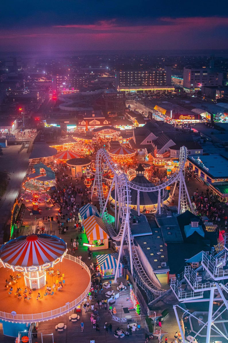 An aerial view of an amusement park at night. The park is lit up with colorful lights, and there are many rides and attractions visible, including a carousel, a roller coaster, and a Ferris wheel. The park is located on a boardwalk, and there are many people walking around and enjoying the rides. The sky is a dark purple color, and there are some clouds in the distance.