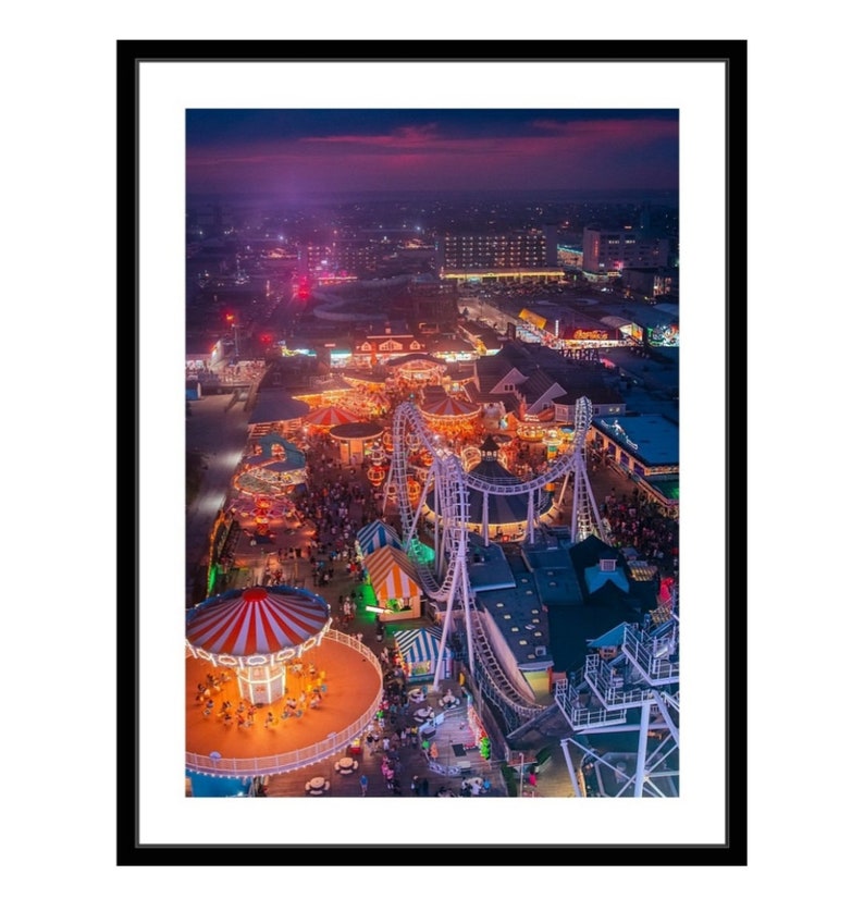 May include: An aerial view of a carnival at night. The carnival is lit up with colorful lights. There are rides, games, and food stalls. The Ferris wheel is in the foreground, and the roller coaster is in the background. The sky is a dark purple color, and the city lights are visible in the distance.
