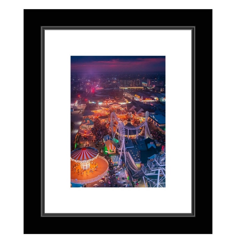 May include: An aerial view of a carnival at night. The image shows a Ferris wheel, a roller coaster, and a carousel. The carnival is lit up with colorful lights. The sky is dark blue with a hint of pink.