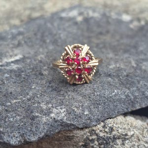 May include: A gold ring featuring a cluster of seven round red gemstones arranged in a circular pattern. The setting includes a twisted rope-like border and four radiating metal prongs extending from the centre to the outer edge of the design.