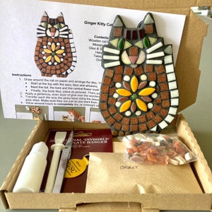 May include: A craft kit for making a mosaic ginger kitty cat. The kit includes a wooden cat base, mosaic tiles, glue, grout, tweezers, and instructions. The kit is packaged in a brown cardboard box.
