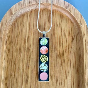 May include: A silver necklace with a rectangular pendant featuring five colourful, circular designs. The pendant's design includes green, pink, yellow, and white elements. The necklace is displayed on a wooden tray.