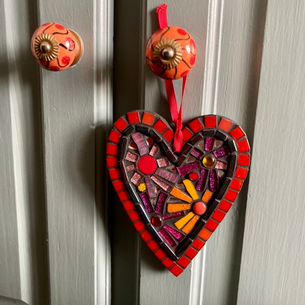 May include: A red heart-shaped mosaic with a black background and colorful glass tiles. The mosaic is hanging from a red ribbon on a gray wooden door.