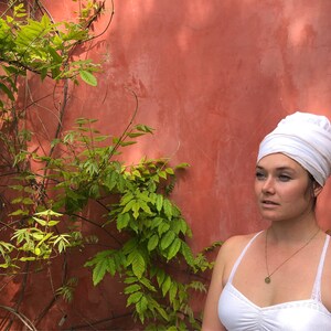 May include: A woman with a white turban on her head, wearing a white tank top and a gold necklace with a pendant. She is standing in front of a red wall with green vines.