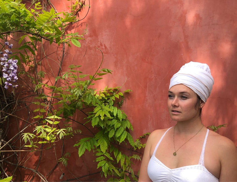 May include: A woman with a white turban on her head, wearing a white tank top and a gold necklace with a pendant. She is standing in front of a red wall with green vines.