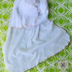 May include: White cotton scarf with a gold trim. The scarf is folded and laid out on a green and white patterned fabric. A measuring tape is placed along the edge of the scarf.
