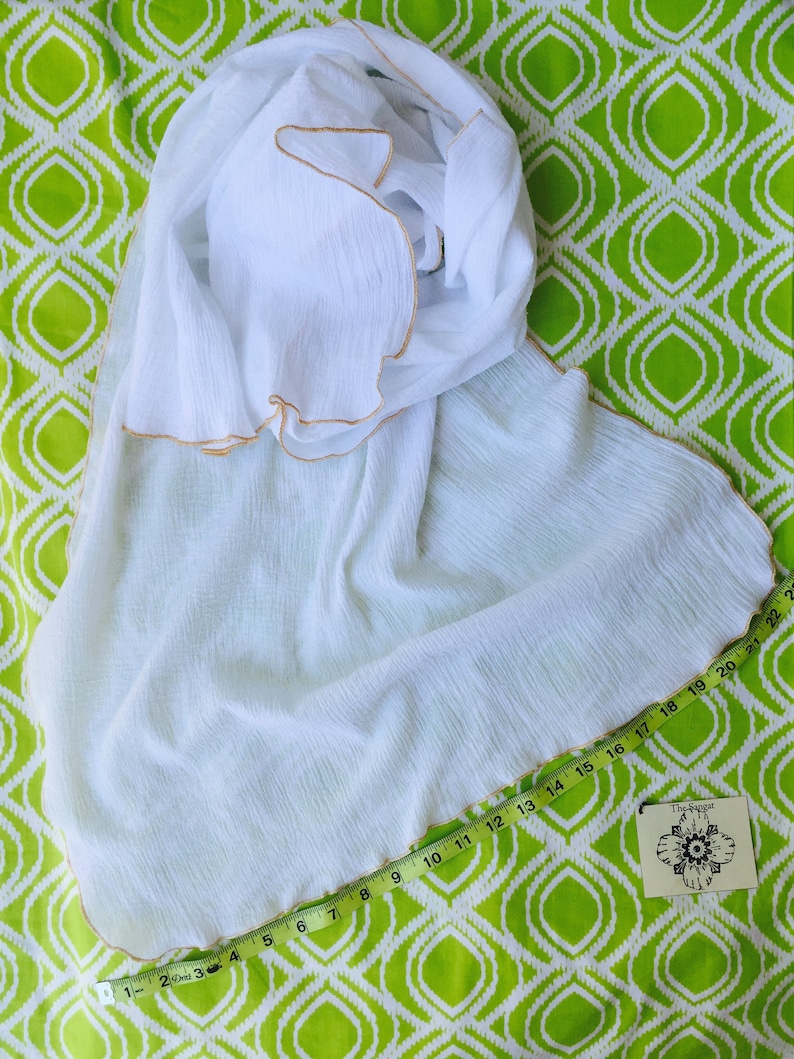 May include: White cotton scarf with a gold trim. The scarf is folded and laid out on a green and white patterned fabric. A measuring tape is placed along the edge of the scarf.