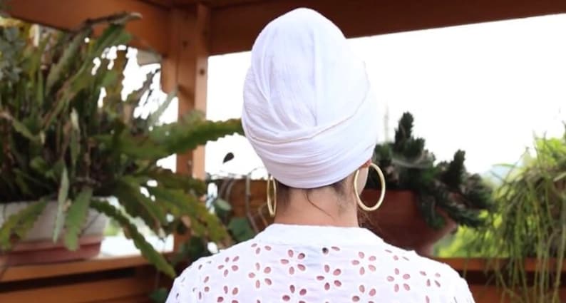 May include: A white turban with a gold trim, worn by a person with gold hoop earrings. The person is wearing a white, lacy top and is standing in front of a green plant.