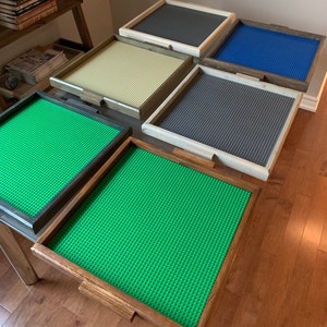 Puede incluir: Una colección de bases Lego con marco de madera en varios colores, incluyendo verde, azul, beige y gris. Cada base tiene una superficie texturizada con pequeños pivotes circulares para fijar los ladrillos Lego. Los marcos tienen un acabado de madera natural.