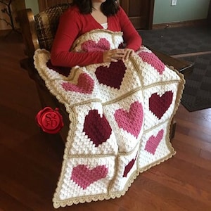 May include: A crocheted blanket with a heart pattern in shades of pink, red, and white. The blanket is made with a white yarn and has a brown border.