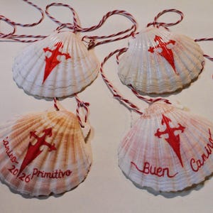 May include: Four white scallop shells, each featuring a painted red Cross of Saint James. Two shells are plain, while the other two are inscribed with the red text Camino Primitivo 2026 and Buen Camino. Each shell hangs from a red and white string.