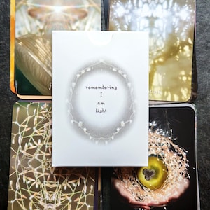 May include: A deck of oracle cards with a white box in the center. The box reads "remembering I am light" in a circular design. The cards have various images, including a glowing apple, lights, and abstract art. The cards are arranged on a dark surface.