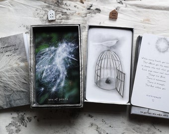 May include: A tarot card deck with a soft, ethereal aesthetic. The deck includes cards with illustrations of a bird on a cage, a dandelion, and text. The box and cards have a muted colour palette, with shades of grey, white, and green.