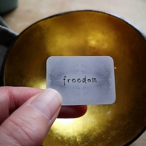 May include: A small card with the word "freedom" written on it is held over a gold bowl. The bowl has a dark rim. The background is blurred. The card is held by a hand.