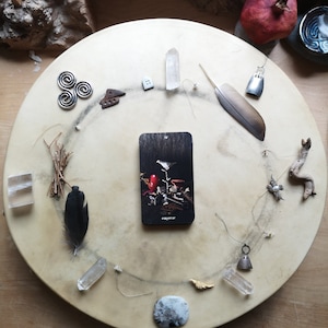 Pu&ograve; includere: Una disposizione circolare di oggetti spirituali su una superficie marrone chiaro. Include cristalli, piume, una carta dei tarocchi, piccoli ciondoli di metallo e un melograno. La carta dei tarocchi presenta un uccello e la parola "emperer".